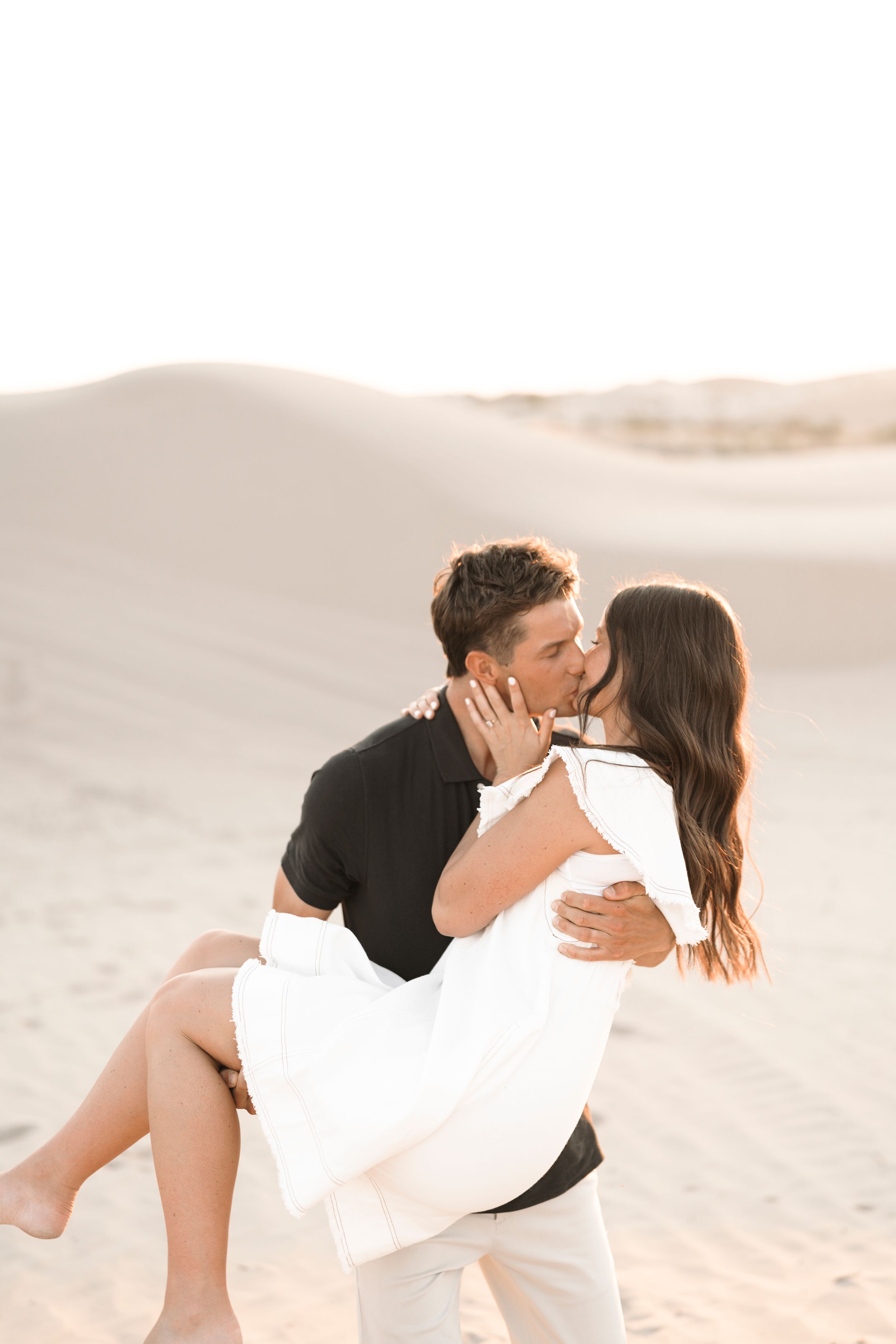 A man in black shirt carries and kisses a woman in white dress on sandy desert dunes.