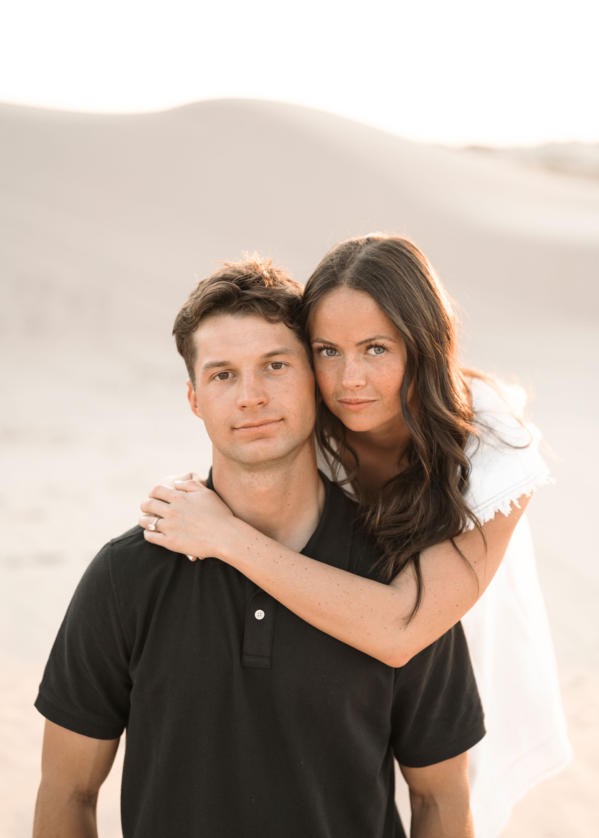 Couple posing together in a sandy desert, woman in white dress embracing man in black polo shirt.