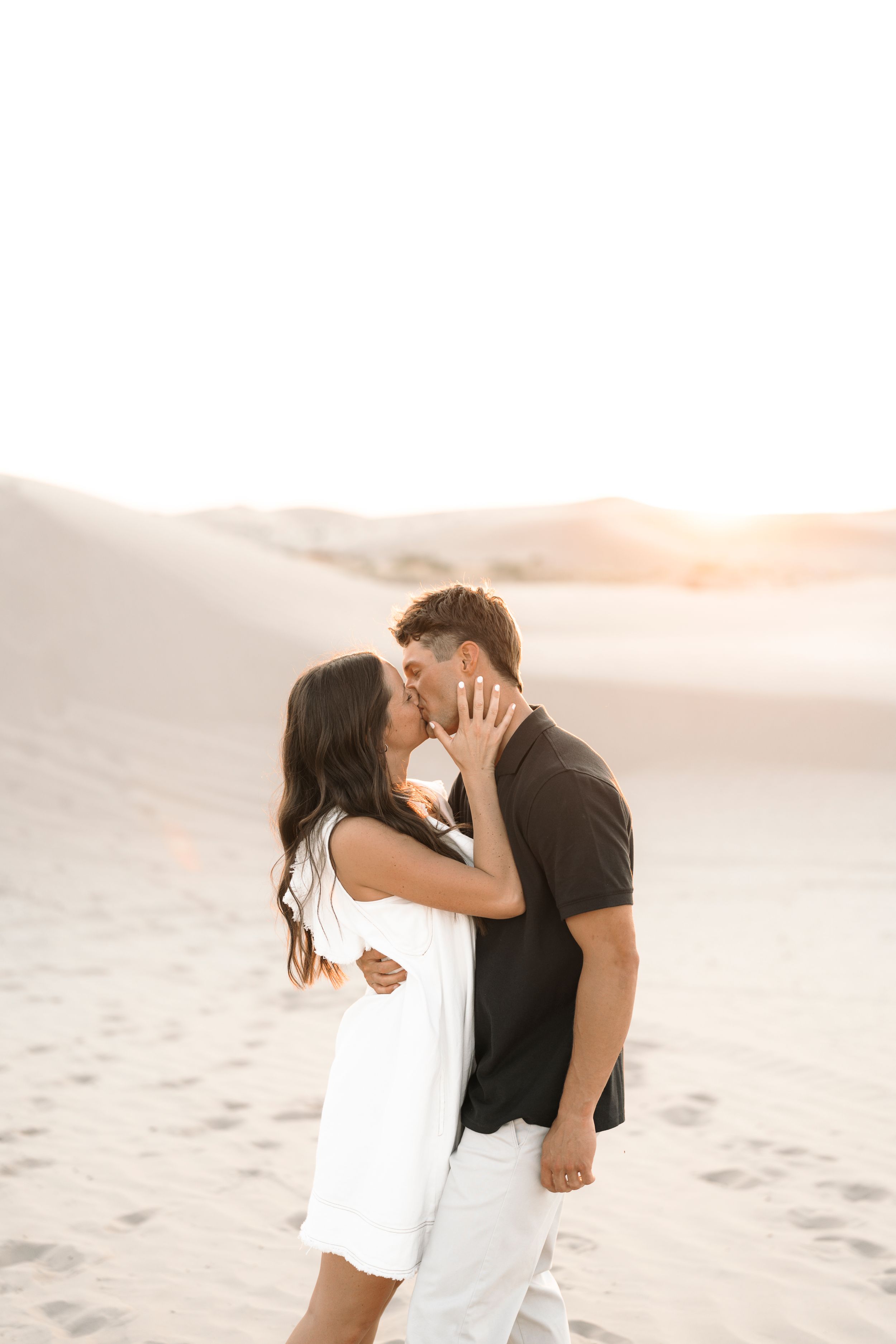 Couple sharing a romantic kiss on sandy dunes at golden hour, woman in white dress, man in black shirt.