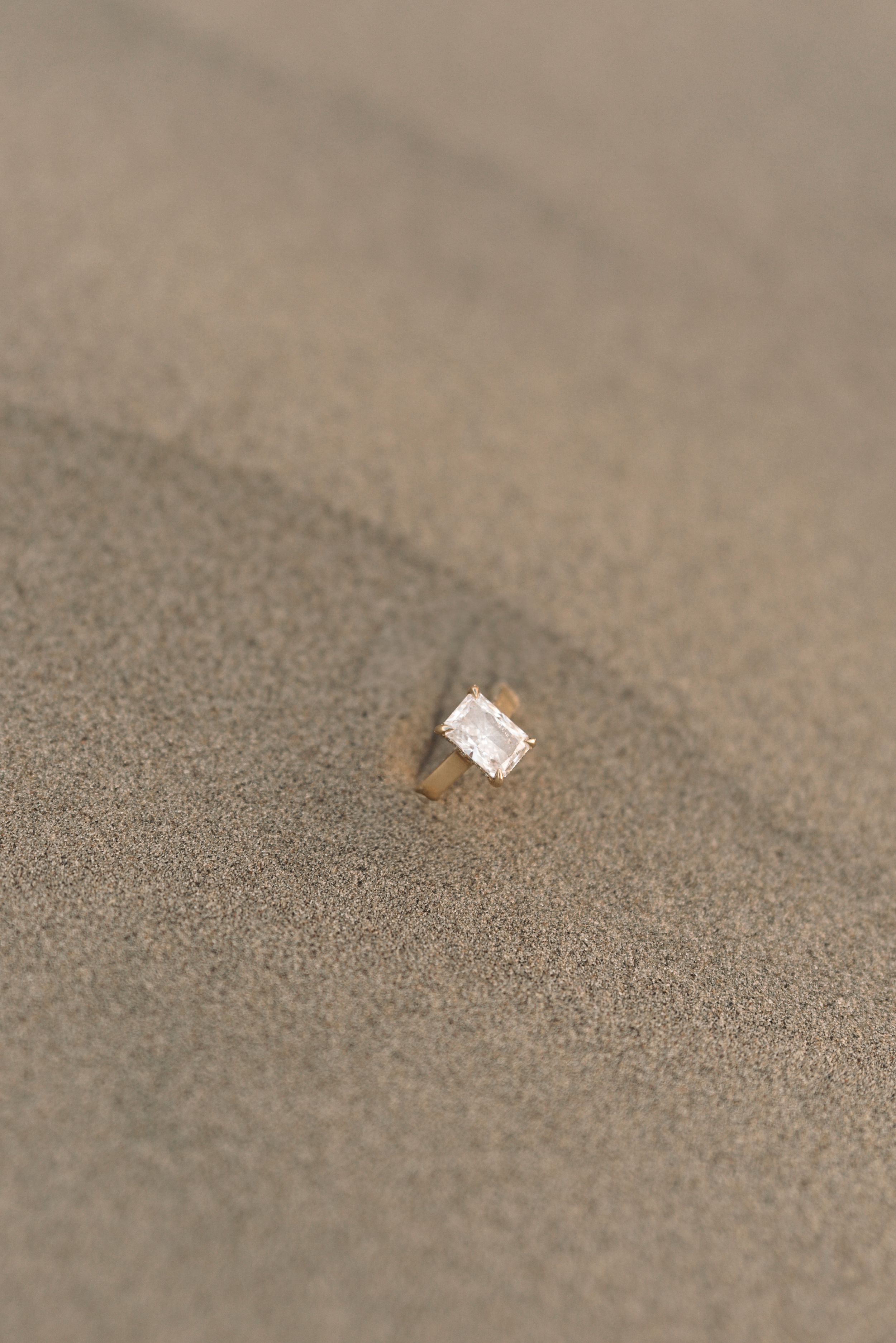 Gold ring with emerald-cut diamond gemstone partially buried in smooth sandy beach surface.