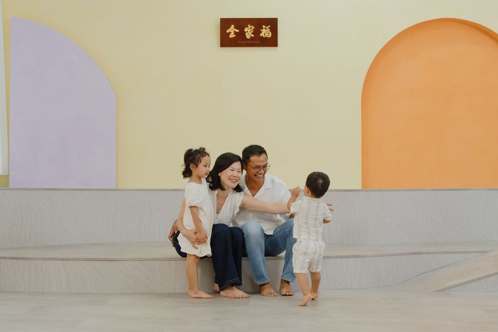 A happy Asian family with two young children playing together in a colorful indoor space with arched walls.