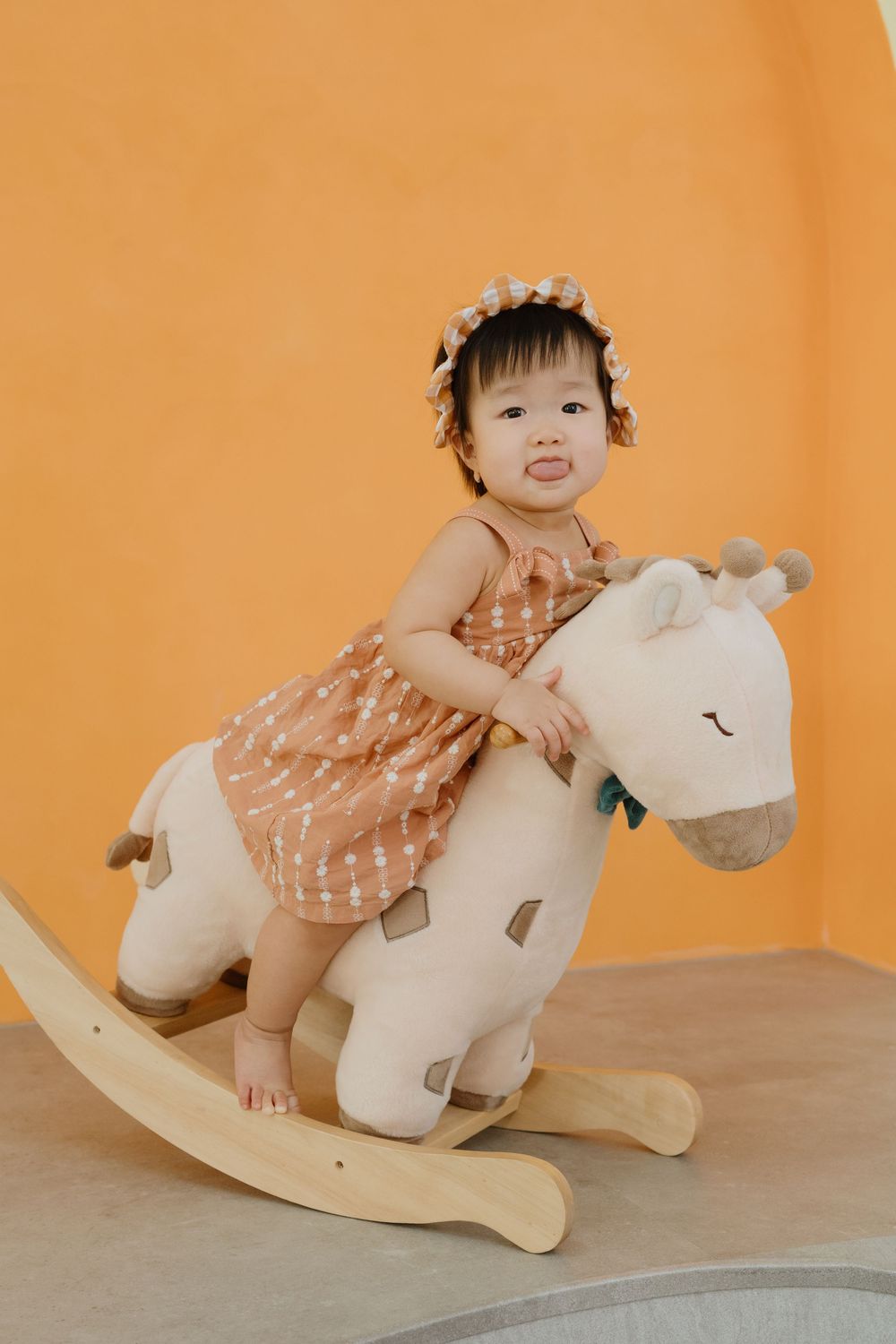 Cute toddler girl in brown polka dot dress riding a white plush rocking horse toy against yellow wall.