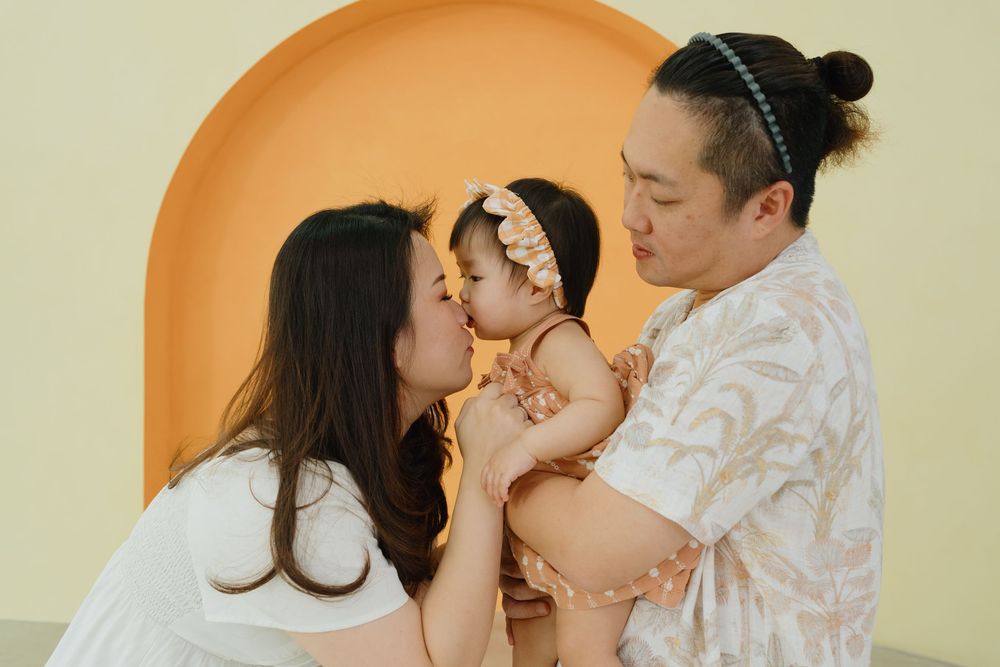 A young Asian family shares a tender moment as parents kiss their baby girl in front of a yellow arch backdrop.