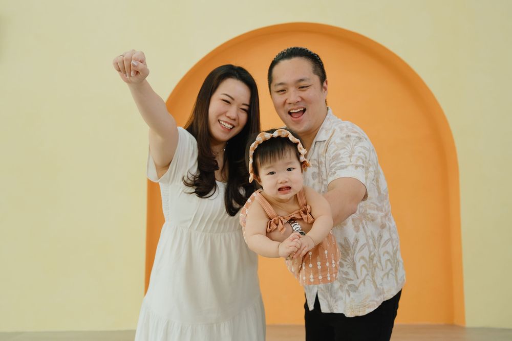 Happy Asian family with parents and baby girl posing in front of yellow arch backdrop.