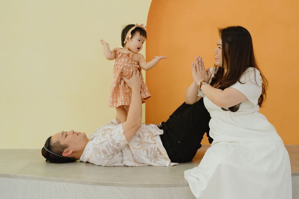 A man lying down lifts a baby girl while a woman in white dress plays with the child indoors.