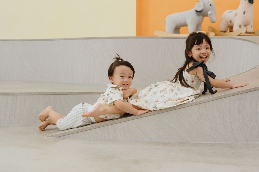 Two young children smiling and sliding down a ramp together in a bright indoor play area.