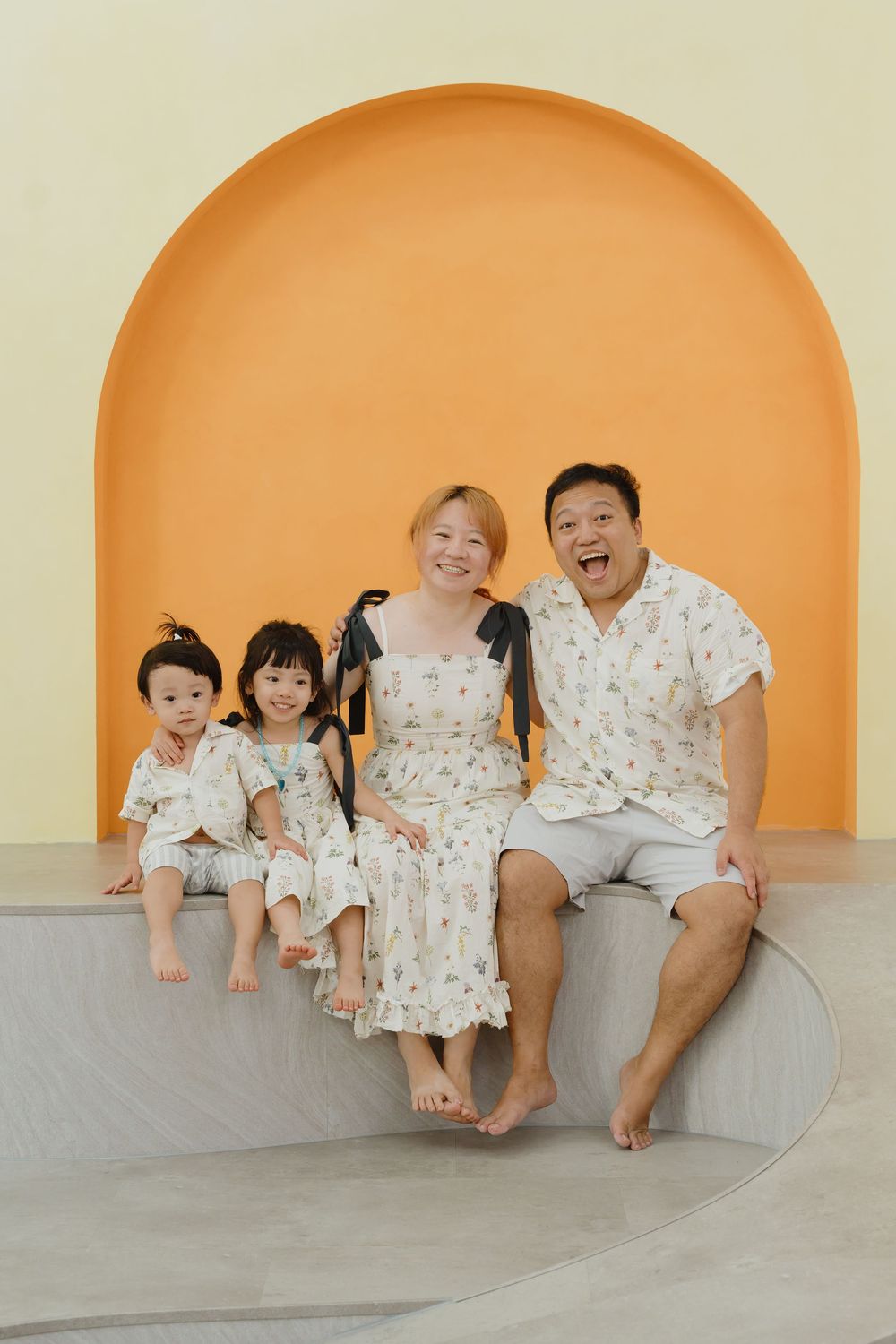 Happy Asian family of four in matching floral outfits sitting together against a warm yellow arched wall backdrop.