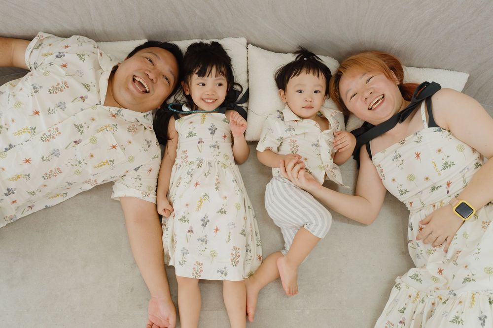 Family of four lying on bed wearing matching floral outfits, two parents with toddler girl and baby boy smiling overhead.