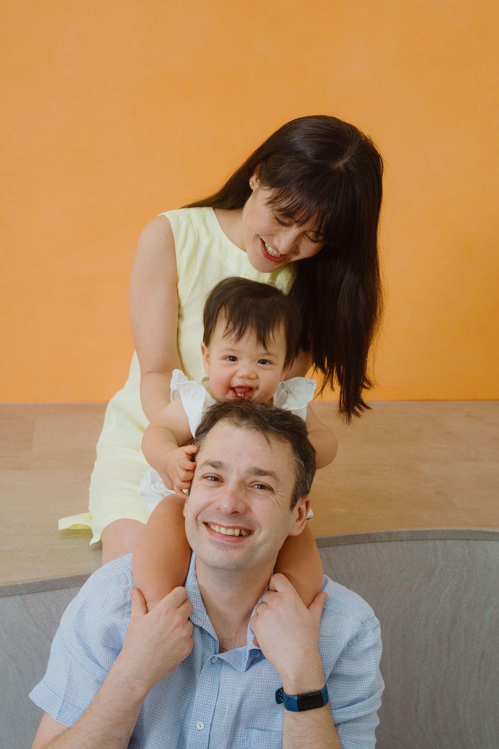 Happy family portrait with smiling father, laughing baby on shoulders, and mother in yellow dress against orange wall.