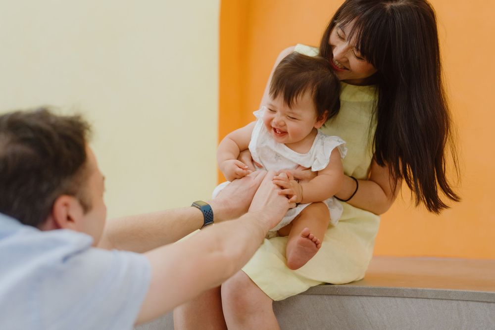 A mother holds a laughing baby while a man reaches out to the child in a warm family moment.