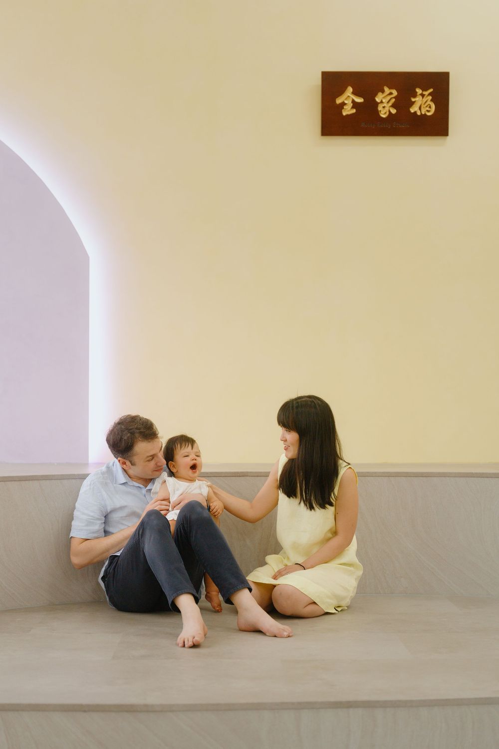 A happy interracial family sits on steps beneath a Chinese blessing sign, parents playing with their laughing baby.