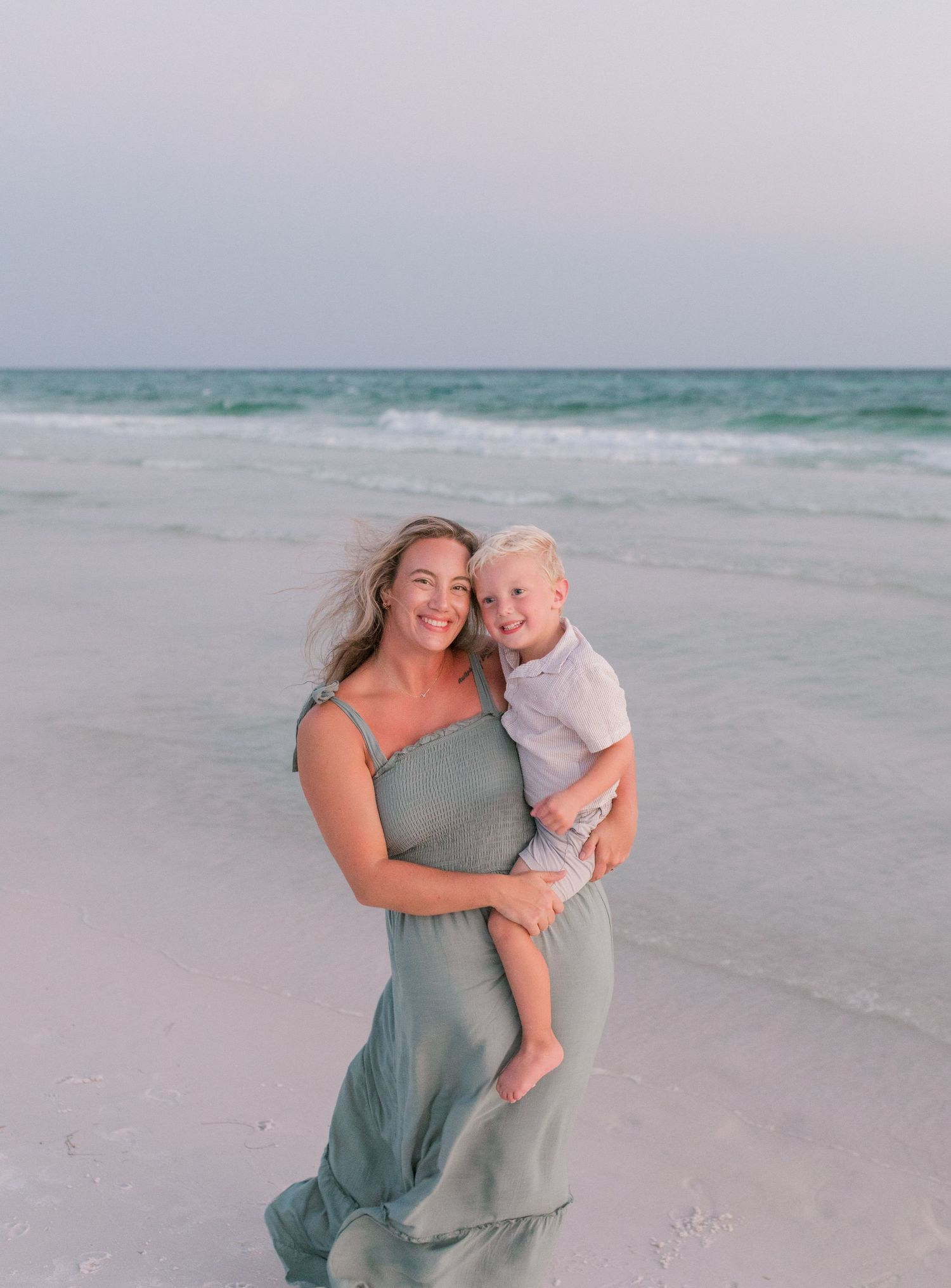 Mother in flowing dress holds child while standing on beach at sunset.