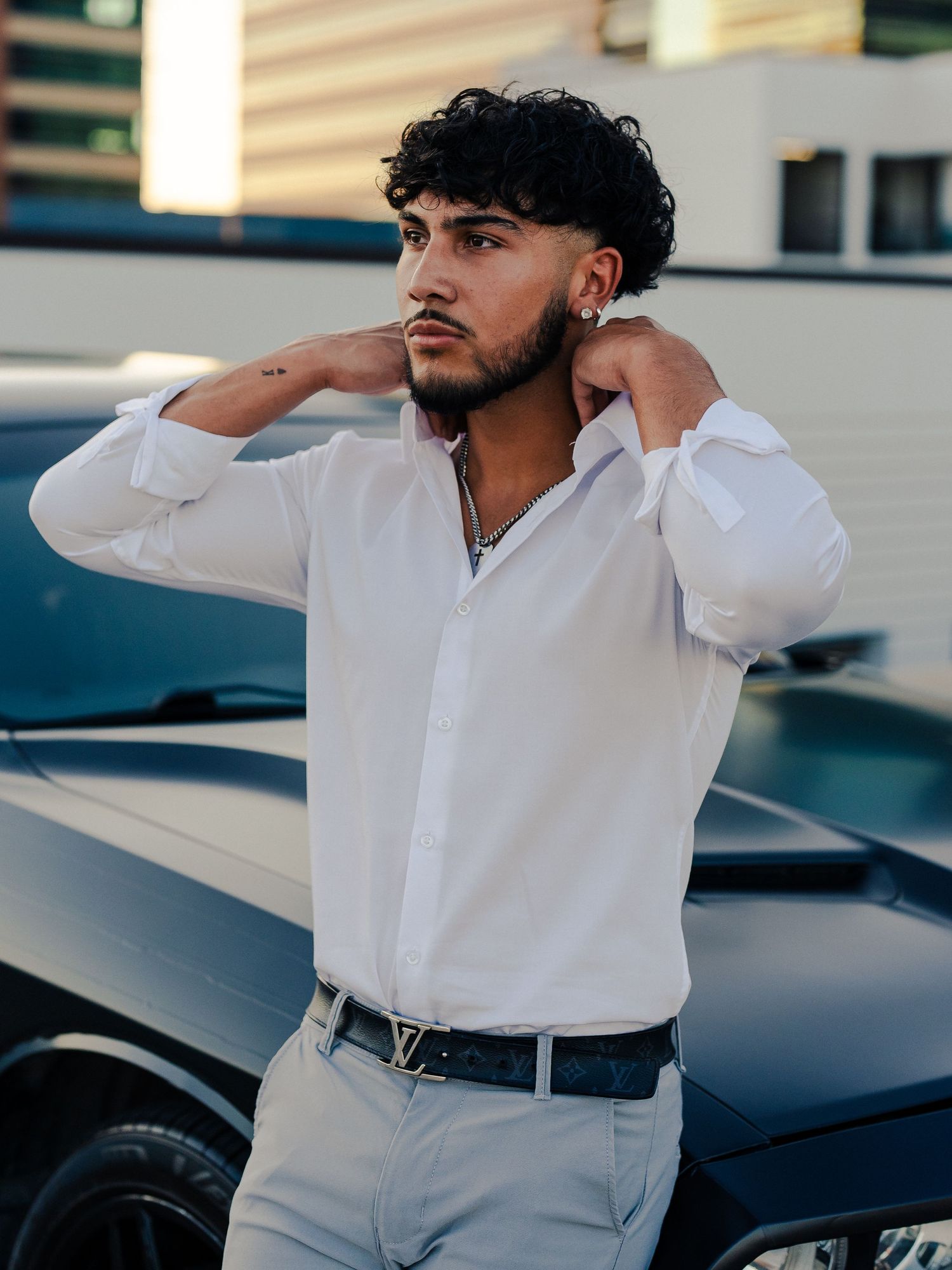 Young man in white button-down shirt poses against car in urban setting.