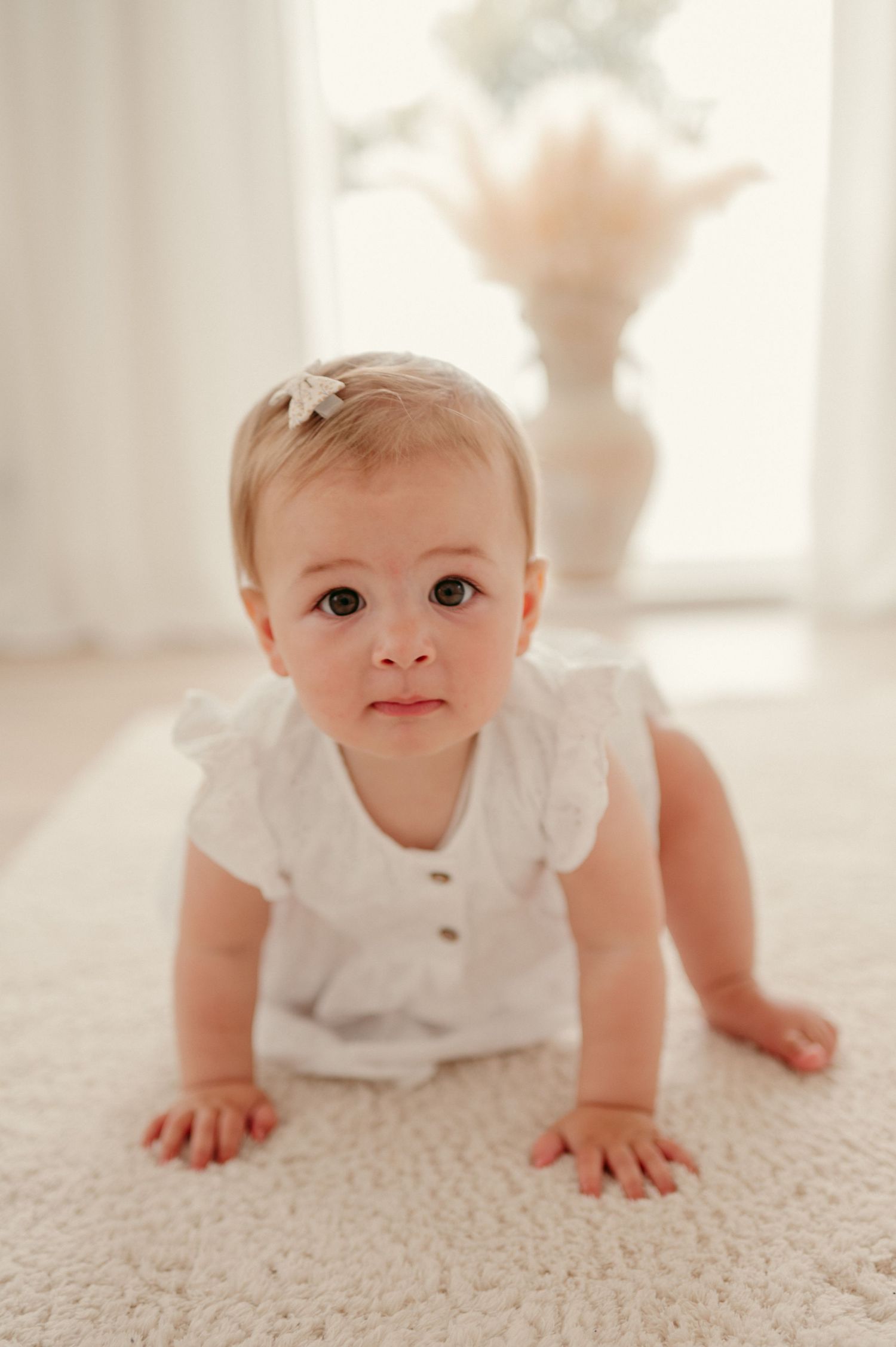 Baby in white clothing crawling on a light carpet in a bright sunlit room.
