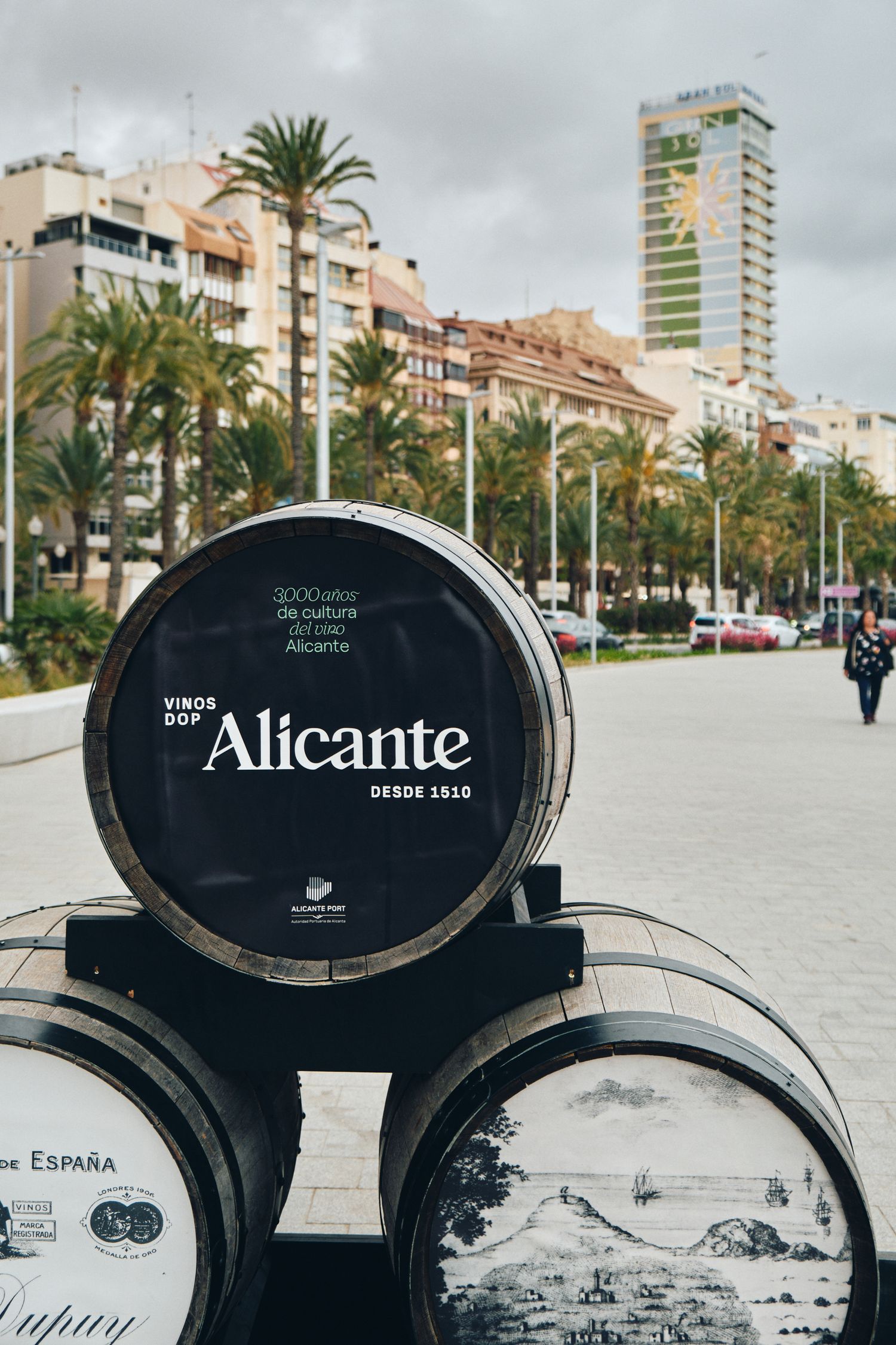 Alicante branded wine barrels displayed along a palm-lined Mediterranean boulevard.