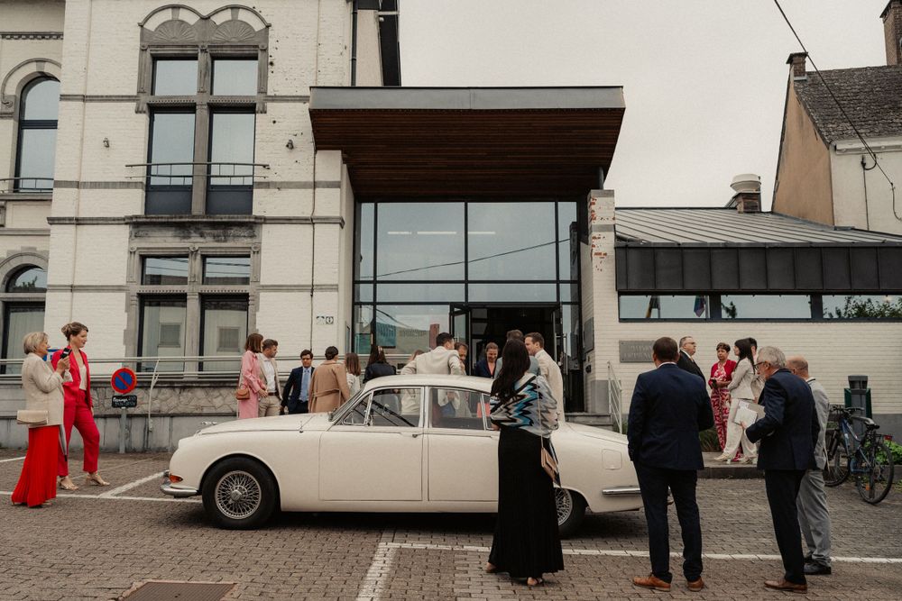Classic white vintage car parked outside historic building with wedding guests gathering around.