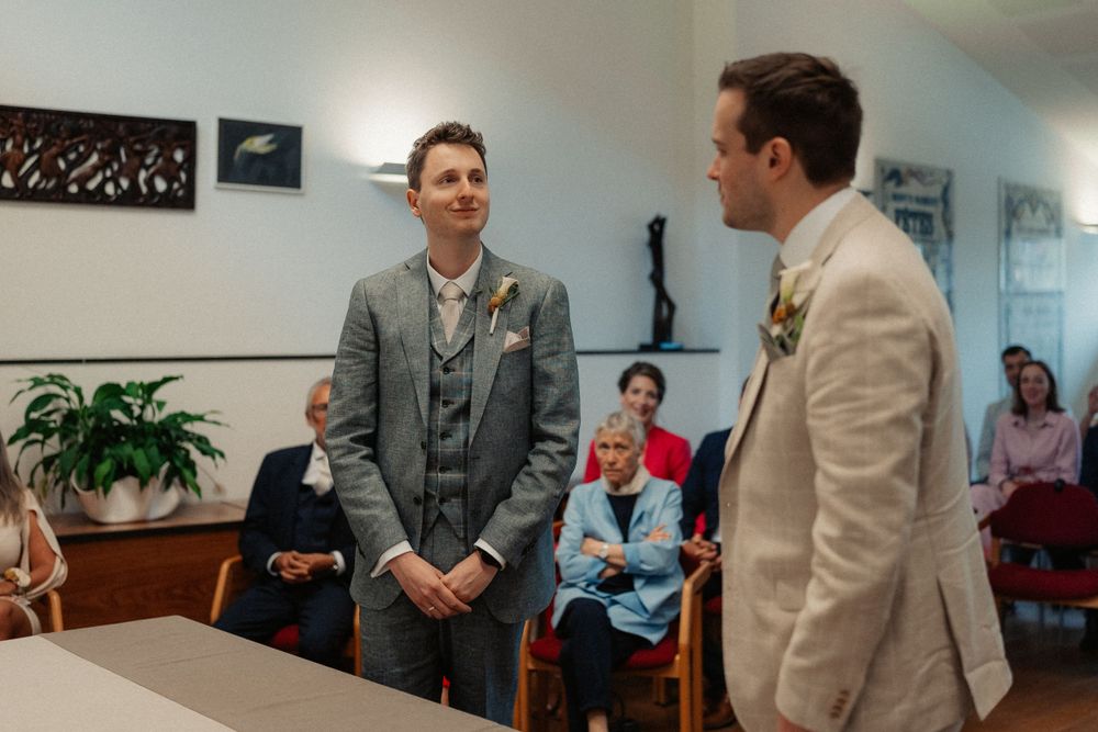 Two people in formal attire standing in a room with indoor plants and waiting guests.