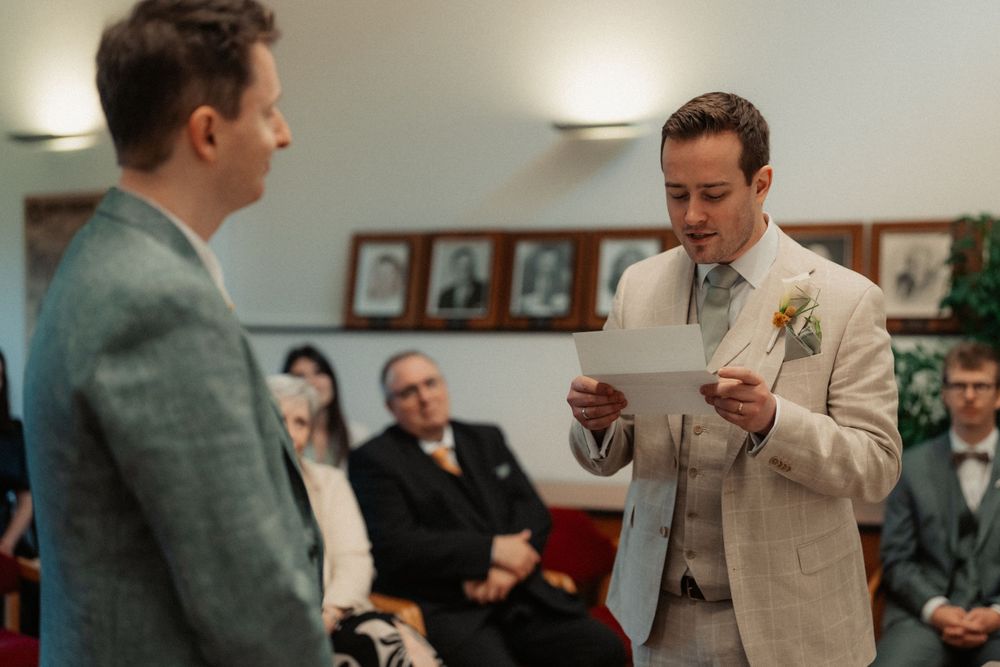 Two people in suits exchange documents during what appears to be a civil ceremony in an office setting.