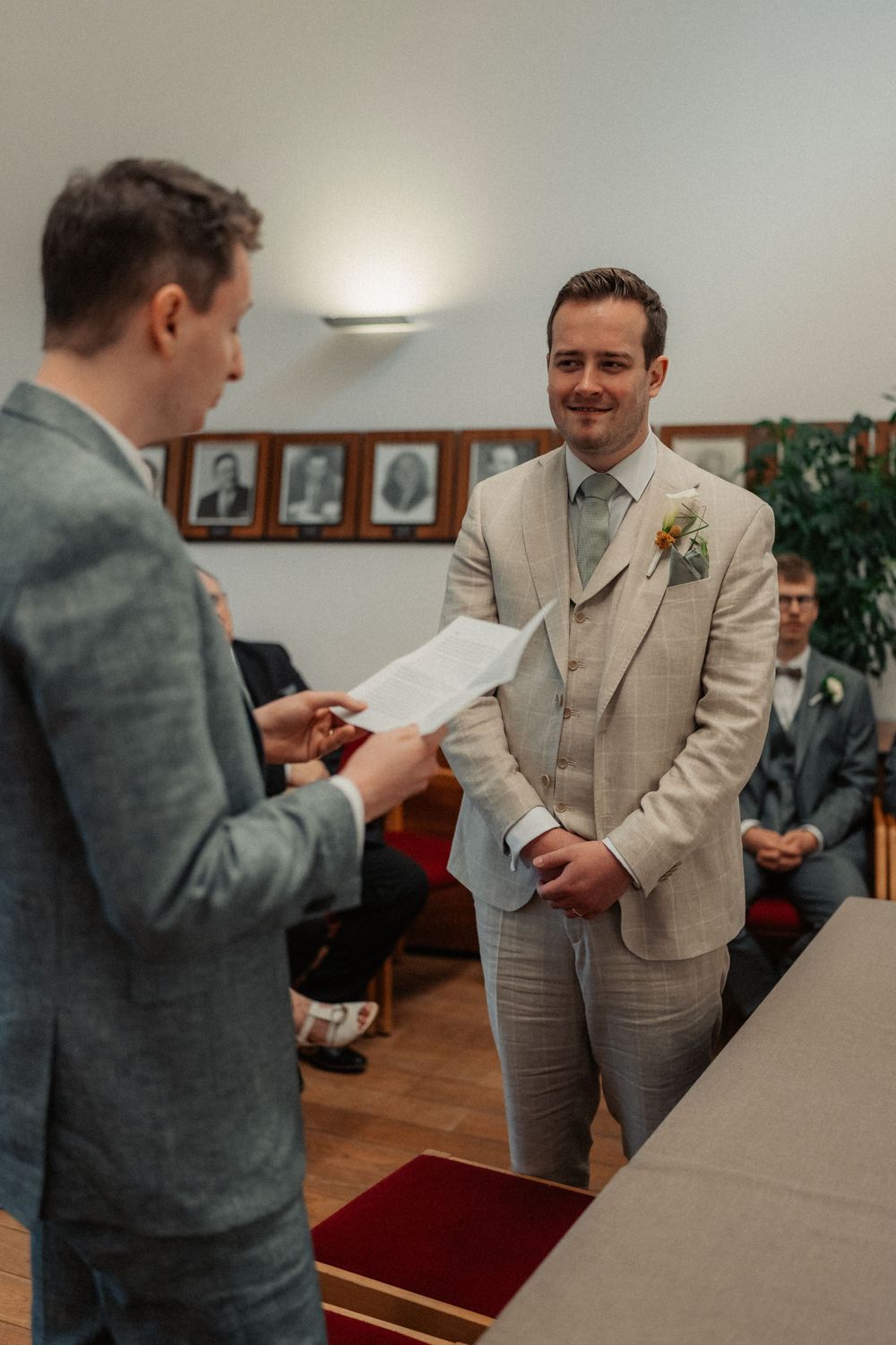A person in a beige suit smiles while another in grey reviews paperwork during a ceremony.