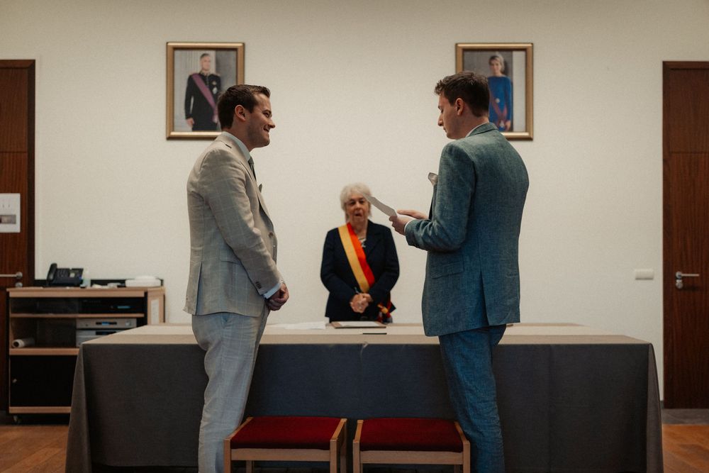 Three people stand in an office room with framed photos and a desk during what appears to be a ceremony.