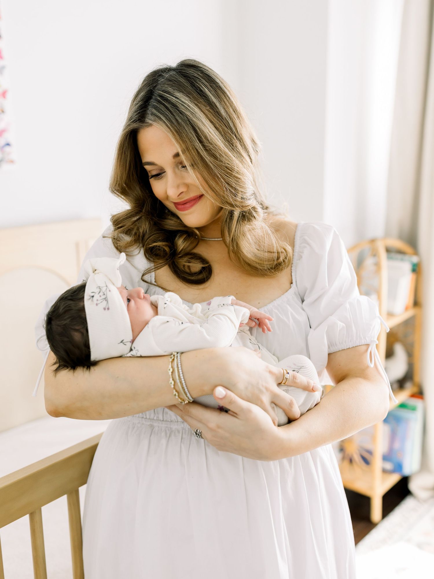 A woman in a white dress tenderly holds a newborn baby in a bright nursery setting.