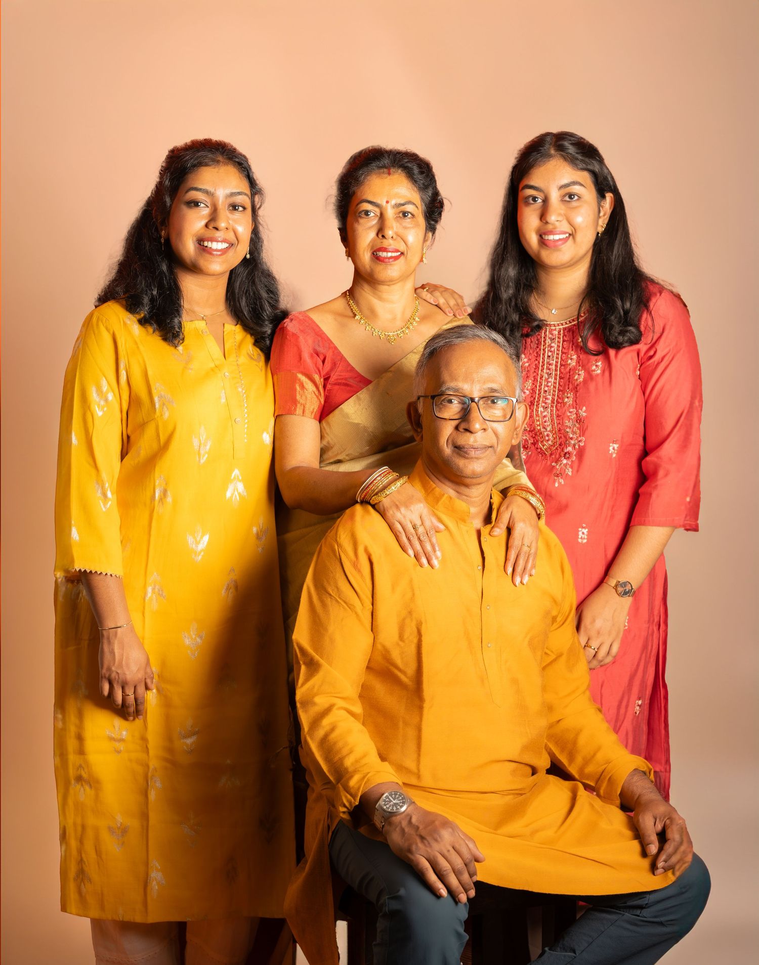 Family of four wearing traditional Indian attire poses together against peach backdrop.