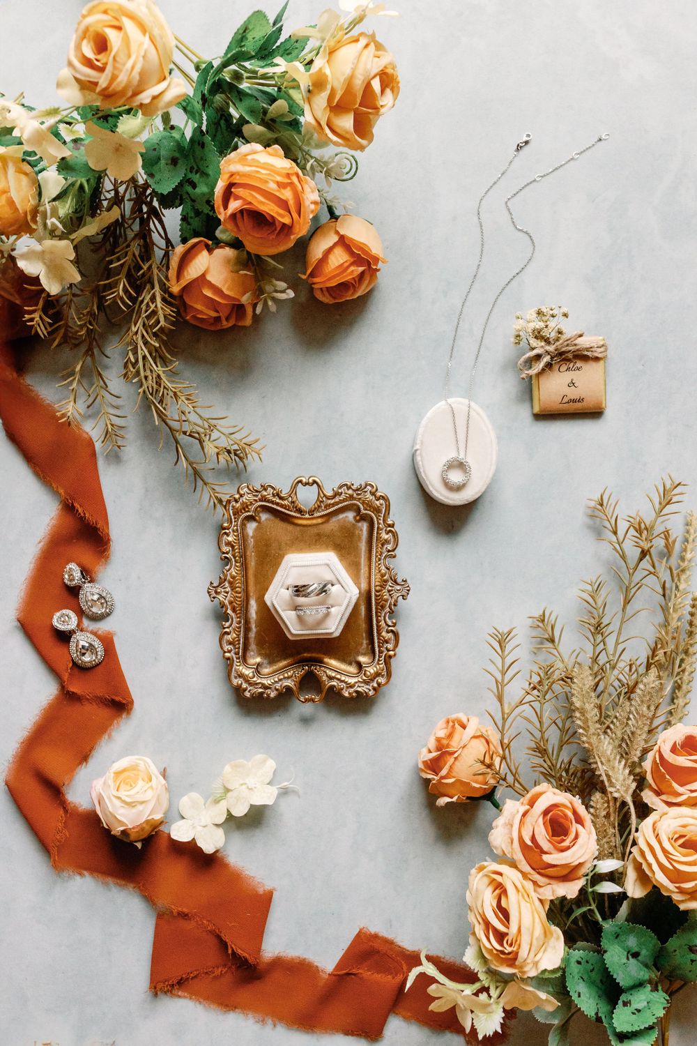 Elegant wedding flat lay with gold-framed ring box, orange roses, rust ribbon, pearl necklace, and earrings on marble surface.