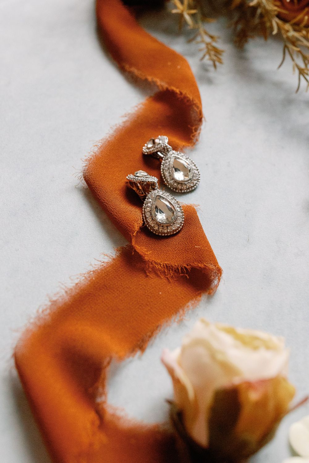 Elegant teardrop crystal earrings resting on a burnt orange ribbon with a blush rose and dried florals on marble.