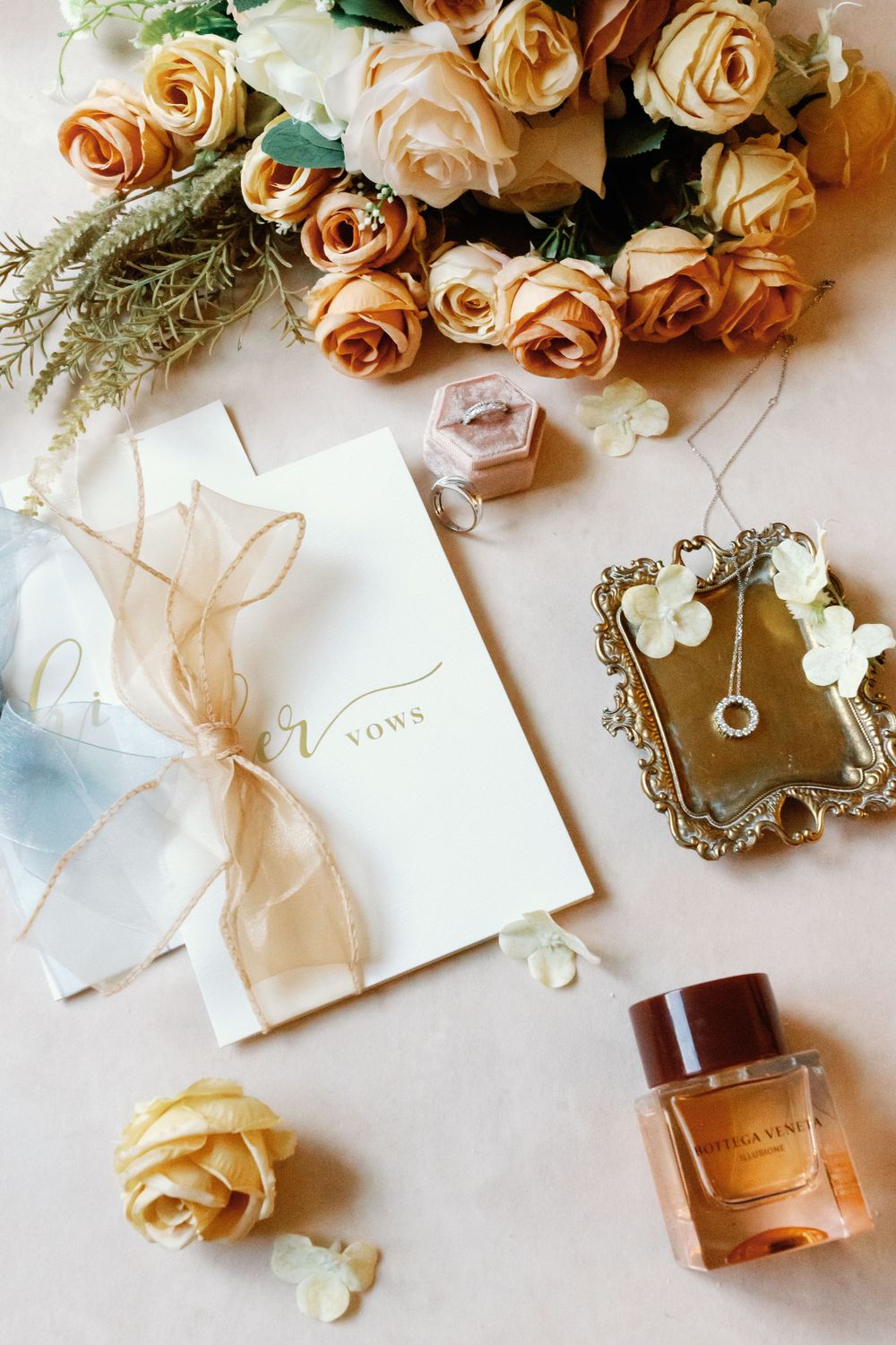 Elegant wedding flat lay with vows booklet, peach roses, diamond necklace, ring box, and Bottega Veneta perfume.