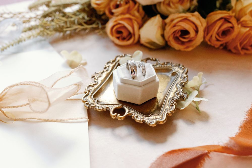 Wedding rings in a white box on an ornate gold tray, surrounded by orange roses and ribbon accents.
