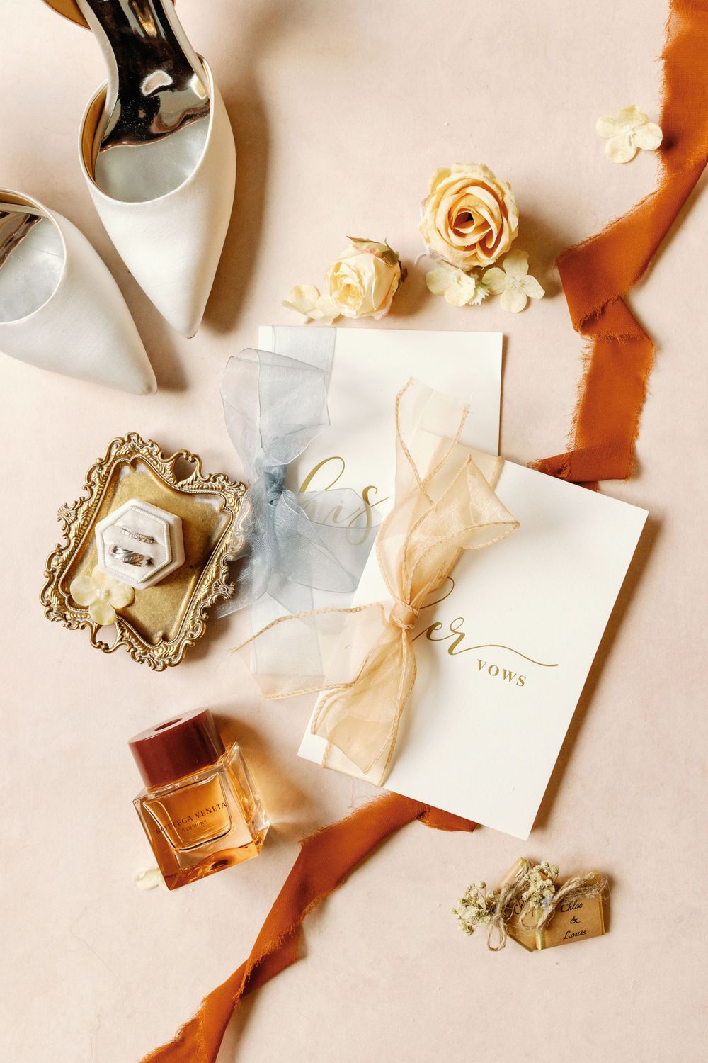 Elegant wedding invitation flatlay with white envelopes, rust ribbons, dried roses, vintage frame, and candle on white surface.