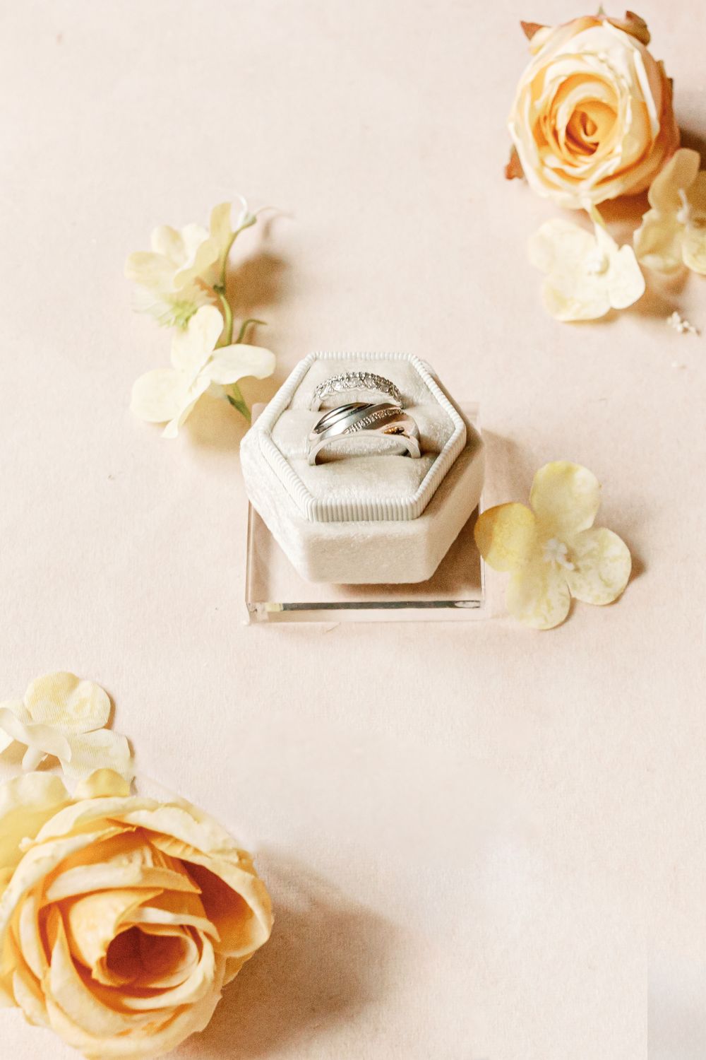 Wedding rings displayed in hexagonal glass box surrounded by cream colored roses and white hydrangea petals on neutral background.