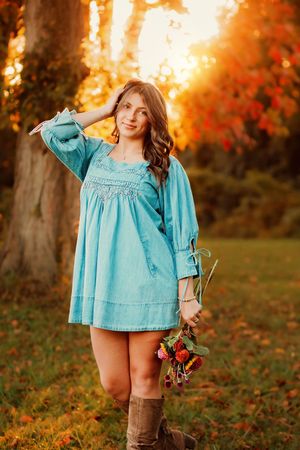 Young woman in teal denim dress holding flowers, posing near a tree during vibrant autumn sunset. Hanover PA Fall Senior Pictures