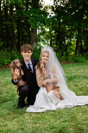 Smiling bride and groom kneel on grass holding two dachshund dogs at outdoor wedding celebration.