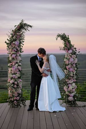 Bride and groom share first kiss under floral arch with pink roses at sunset outdoor wedding ceremony.
