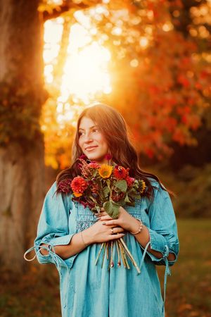 Smiling young woman in blue dress holding colorful autumn bouquet of dahlias in golden sunset light. Hanover PA Fall Senior Pictures