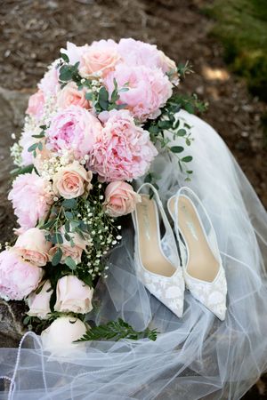 Pink peony and rose bridal bouquet with white lace heels and veil on stone surface.