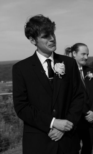 Black and white portrait of groom in dark suit with rose boutonniere at outdoor wedding.