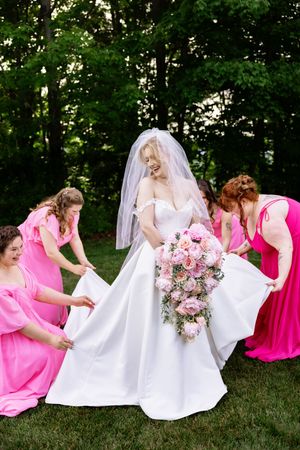 Bride in white gown holds pink bouquet while four bridesmaids in pink dresses arrange her train.