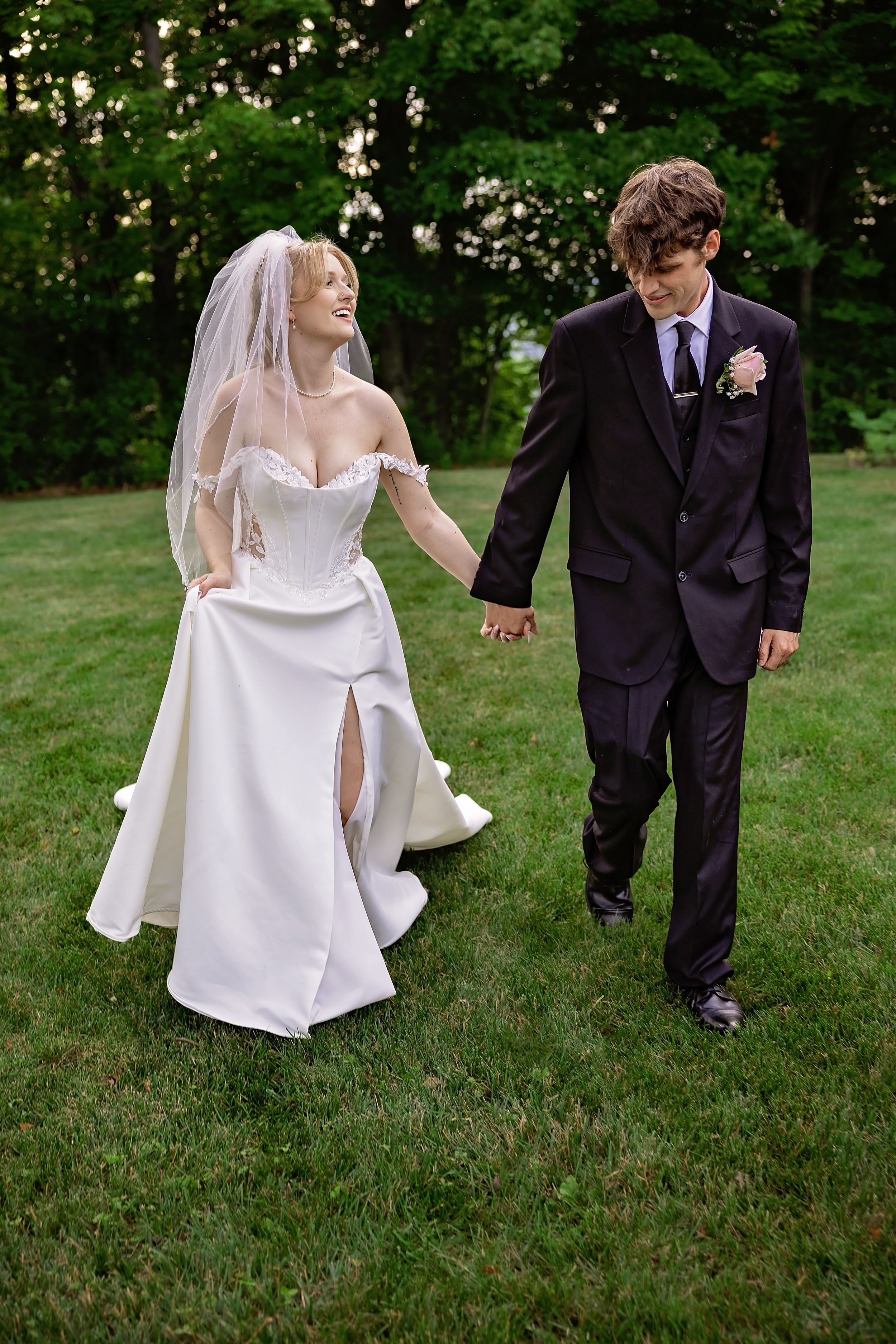 Bride and groom walk hand in hand across green lawn at outdoor summer wedding celebration.