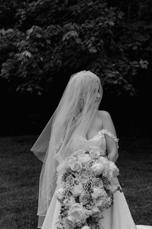 Black and white artistic portrait of bride with veil over face holding large floral bouquet outdoors.