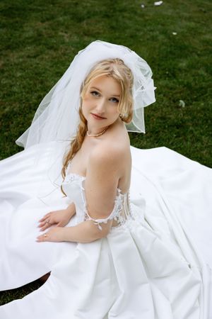 Blonde bride in white lace gown and veil sits on grass looking over shoulder at camera.
