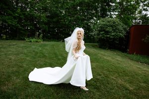 Bride in white wedding gown and veil walks barefoot across lush green lawn outdoors.
