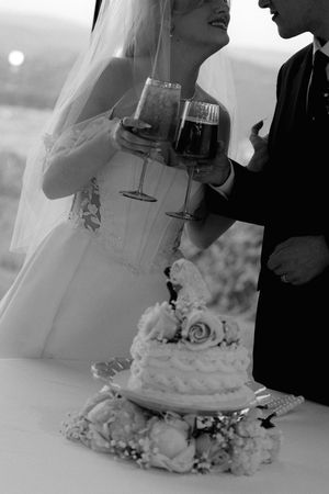 Black and white photo of bride and groom toasting with wine glasses beside floral wedding cake.