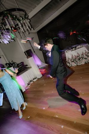 Groom in black suit jumps joyfully on wooden dance floor during lively wedding reception.