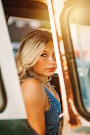 Young blonde woman in denim overalls gazing at camera through a vehicle door in warm sunlight. Hanover PA Spring Senior Pictures
