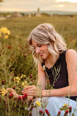 Blonde woman in black top smiling while picking wildflowers in a colorful field at sunset. Gettysburg Senior Pictures