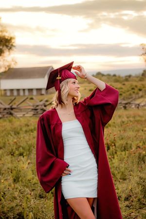 Smiling graduate in maroon cap and gown standing in a field during a golden sunset. Gettysburg Senior Pictures cap and gown