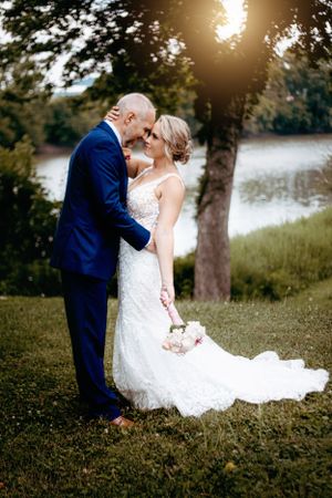 Bride holding bouquet and groom embracing near a sunlit lake on wedding day.