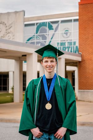 Young male graduate in green cap and gown smiles proudly wearing gold medal outside school.